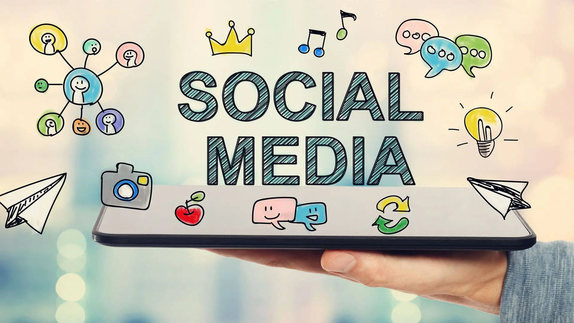 Hand holding a digital tablet displaying bold SOCIAL MEDIA text surrounded by colorful marketing icons and speech bubbles.