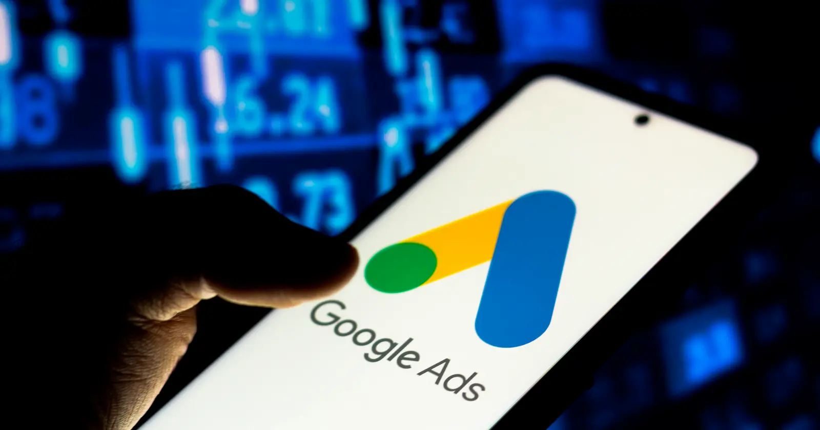 Hand holding a smartphone displaying the Google Ads logo set against a dark background with blurred financial analytics charts.